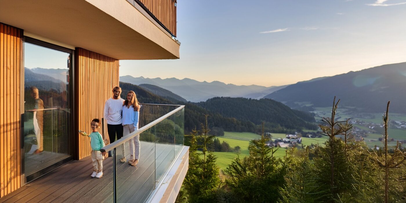 Junge Familie mit Kind auf dem Balkon einer modernen Neubauwohnung mit Holzfassade, Blick auf die Kärntner Berge